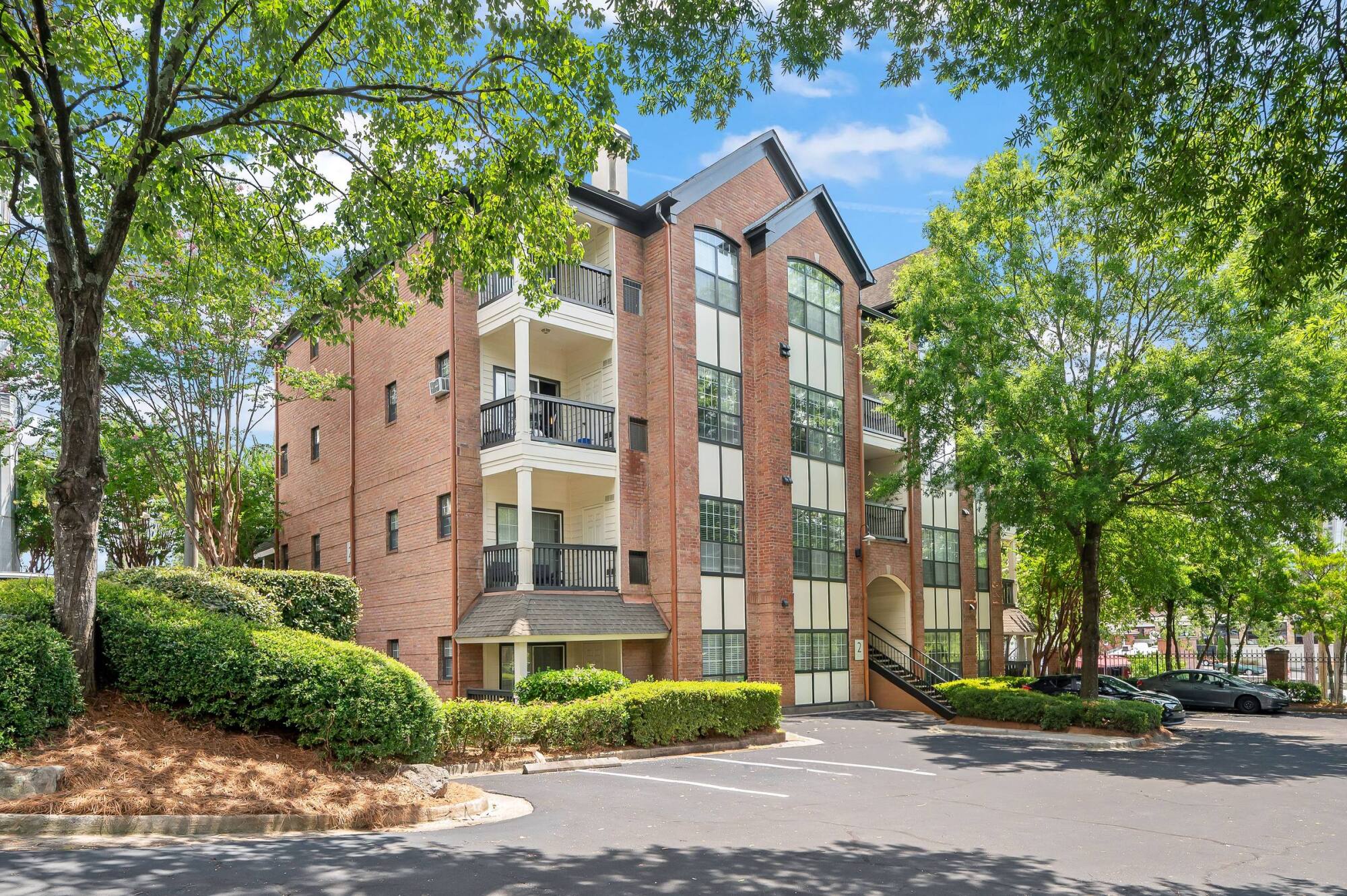 Three-story brick apartment building with balconies, large windows, and surrounding trees on a sunny day.
